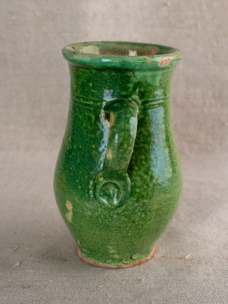 Green glazed French earthenware petite cruche with textured finish and chips, displayed on beige fabric surface.