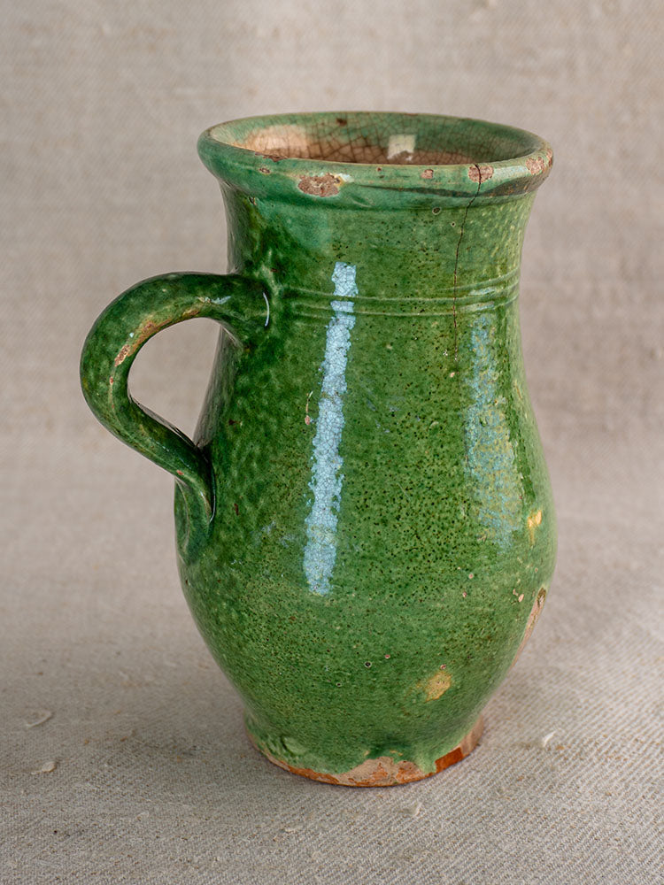 Petite antique French green-glazed earthenware cruche with single handle, chipped rim and base, on beige fabric.
