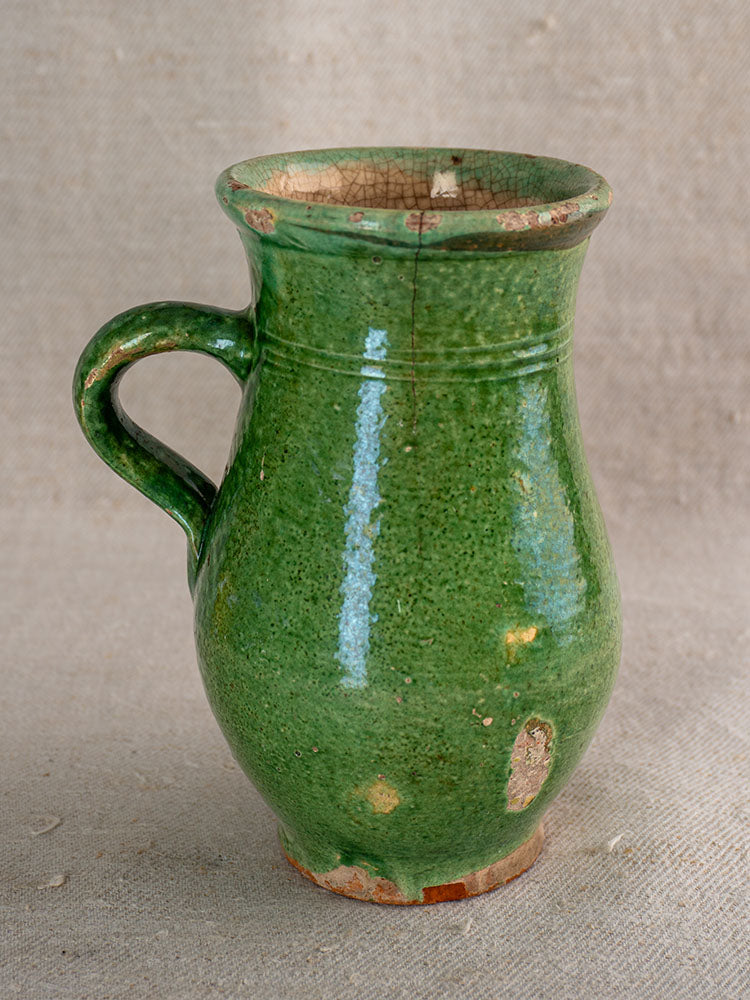Petite antique French green-glazed earthenware cruche with single handle, visible chips, on neutral fabric background.
