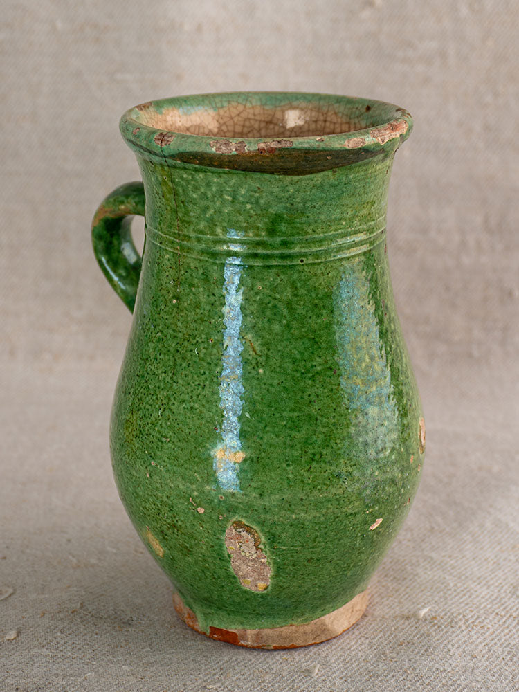 Antique French green glazed earthenware cruche with handle, visible wear, chips, and crackled rim on neutral textured surface.