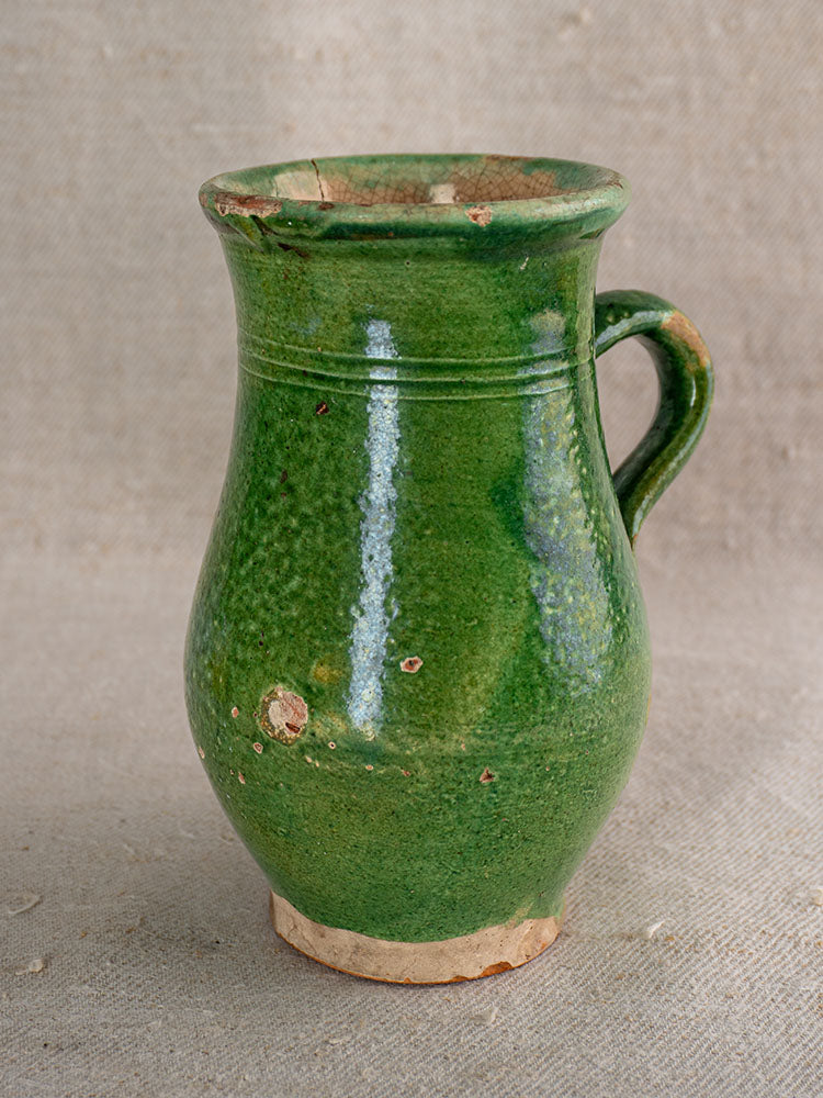 Antique French green glazed earthenware petite cruche with handle, chips and wear, on textured neutral fabric.