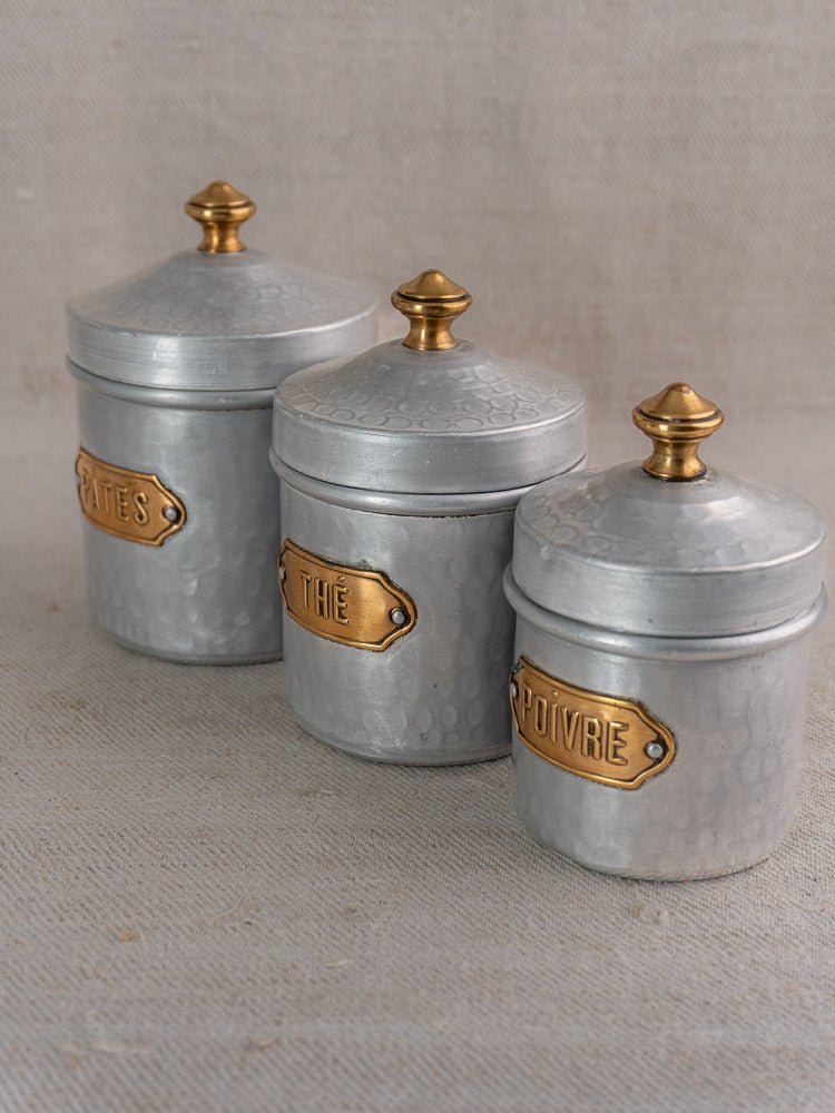 Set of three vintage French hammered aluminum canisters with gold knobs and brass labels.