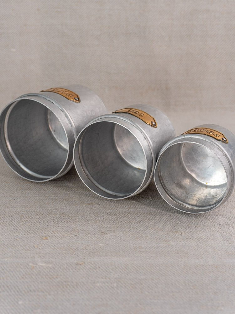 Vintage French hammered aluminum canisters with brass labels, diagonally arranged by size on beige fabric.