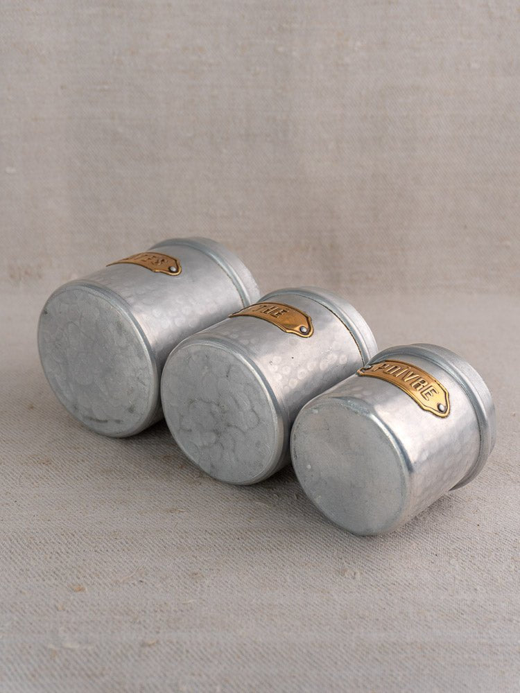 Set of three vintage French hammered aluminum canisters with brass labels on a light beige surface.