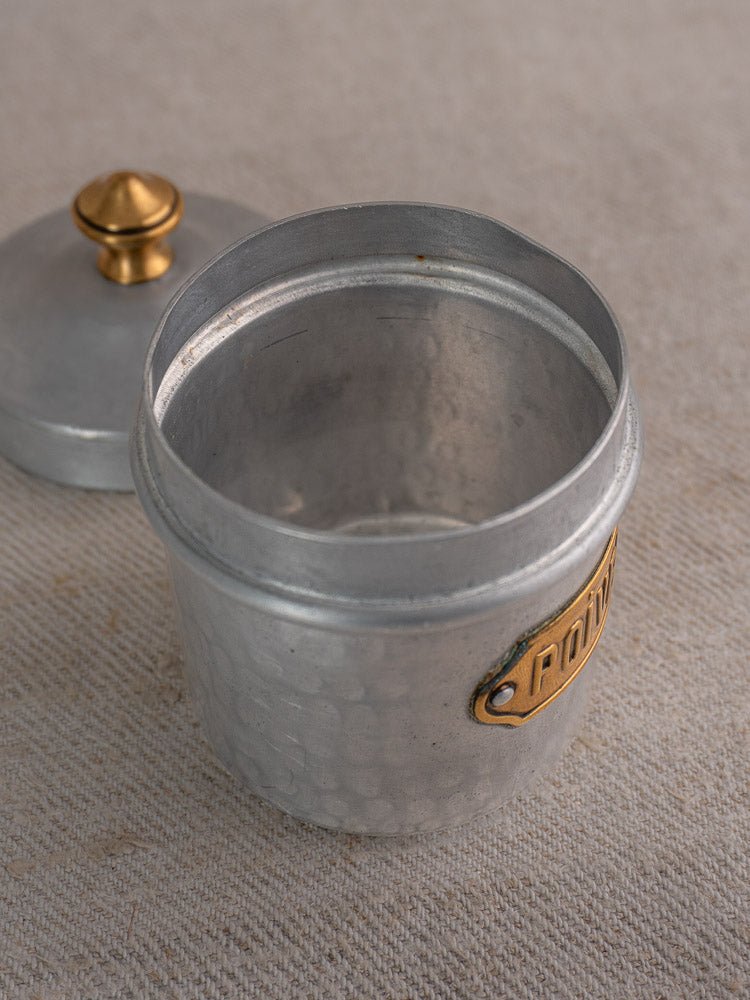Small vintage French hammered aluminum canister with gold knob and brass label on beige textured surface.