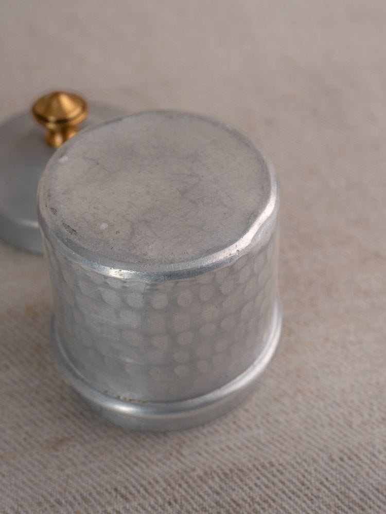 Small round vintage French aluminum canister with hammered texture and brass-accented lid on beige fabric.
