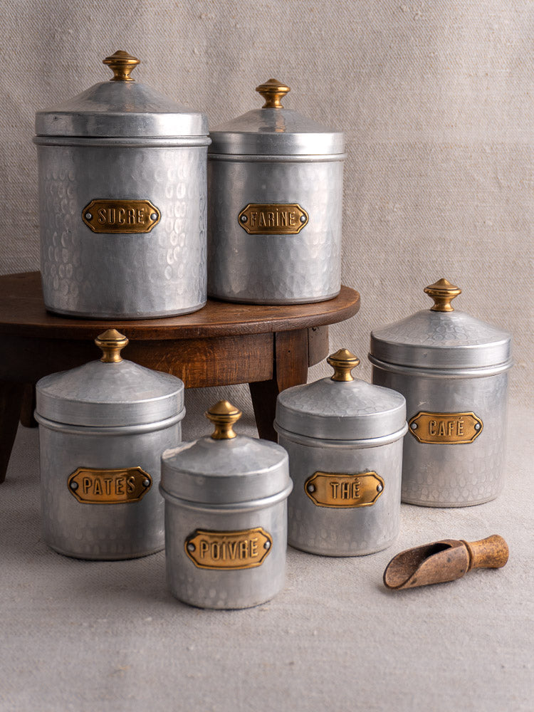 Vintage French hammered aluminum canister set with brass labels and gold knobs on neutral cloth.