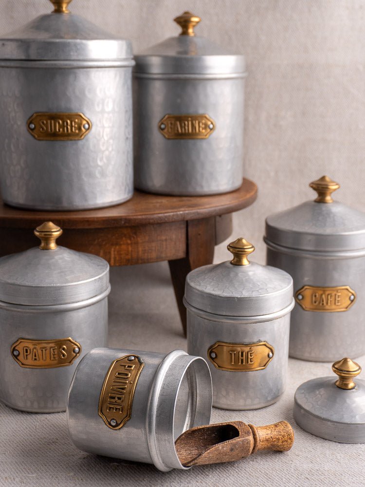 Vintage French hammered aluminum canister set with brass labels on a wooden stand, one jar sideways with scoop.