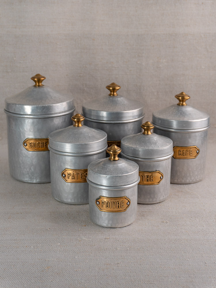 Vintage French hammered aluminum canister set with brass labels and gold knobs on neutral cloth.