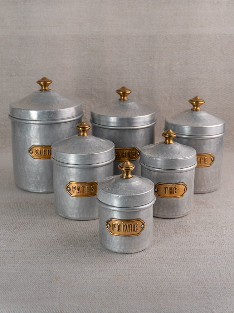 Vintage French hammered aluminum canister set with gold knobs and brass labels on fabric background.