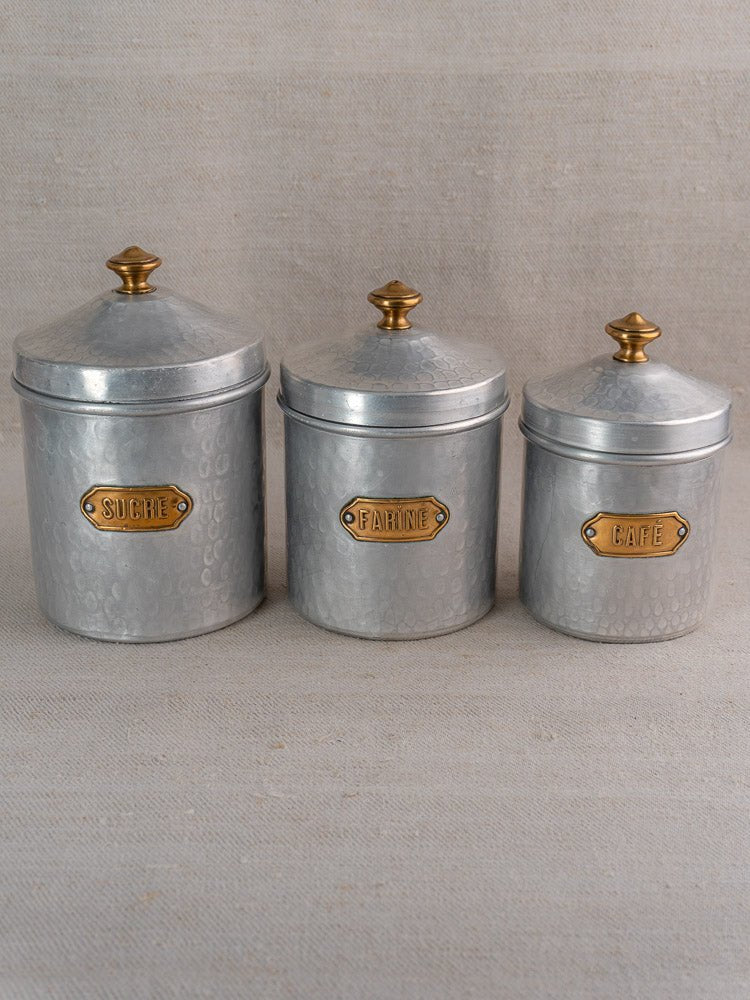 Vintage French hammered aluminum canister set with gold knobs and brass French labels on beige fabric.