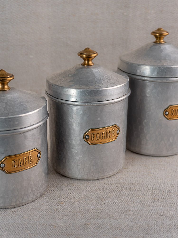 Vintage French hammered aluminum canister set with brass labels and gold knobs on beige fabric.
