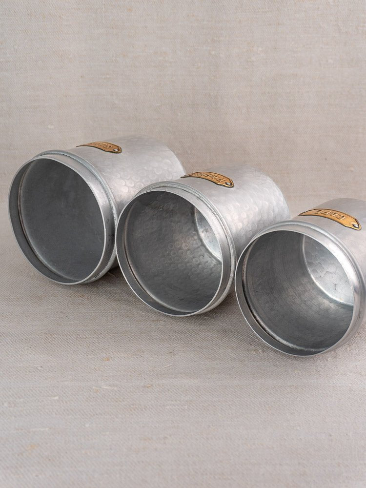 Vintage French aluminum canister set with hammered silver surfaces and brass labels on beige fabric.