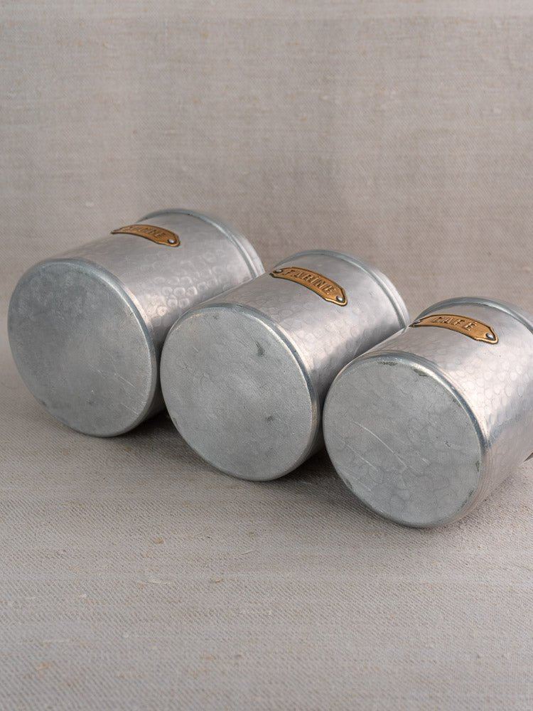 Vintage French aluminum canister set with hammered finish and brass labels on beige fabric surface.