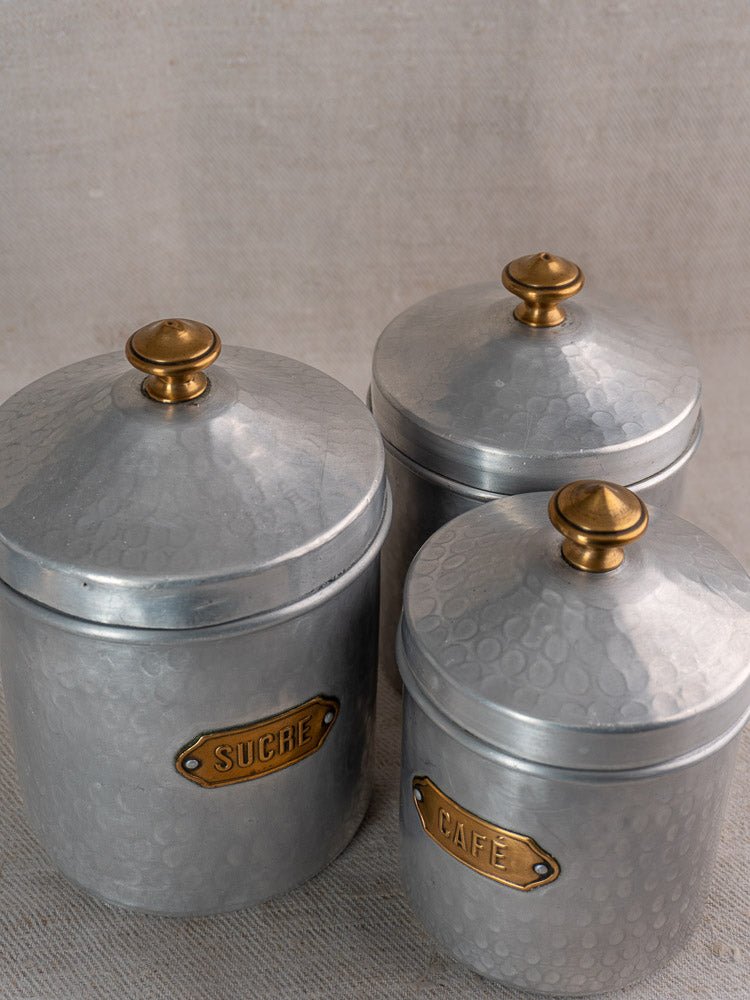 Vintage French hammered aluminum canister set with brass details and SUCRE, CAFE labels on beige surface.