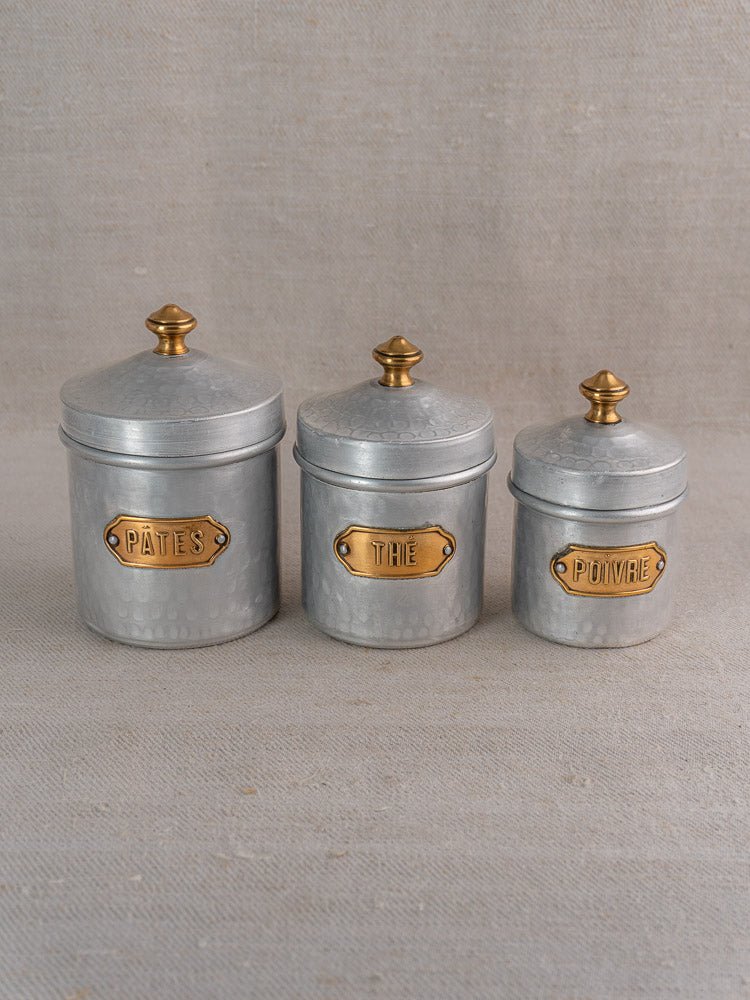 Set of three vintage French hammered aluminum canisters with brass labels and gold knobs on beige fabric.