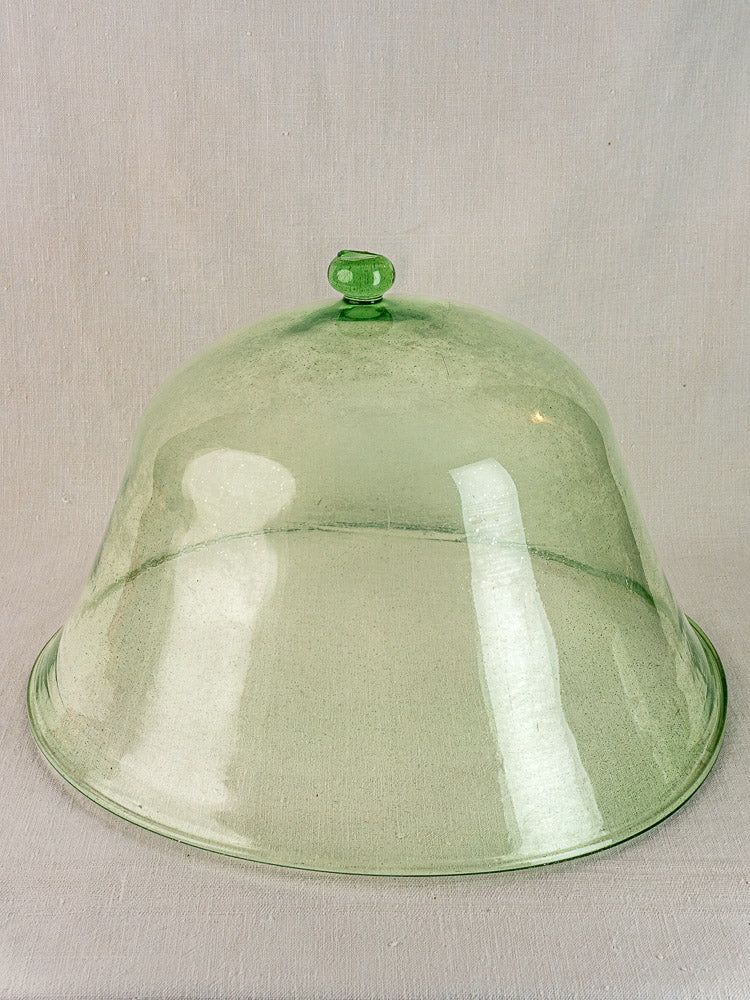 Antique French glass garden cloche, hand-blown pale green with round handle, on light beige fabric background.