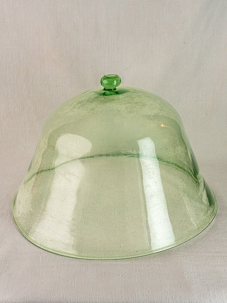 Antique French hand-blown glass garden cloche in pale green with round handle on beige fabric background.