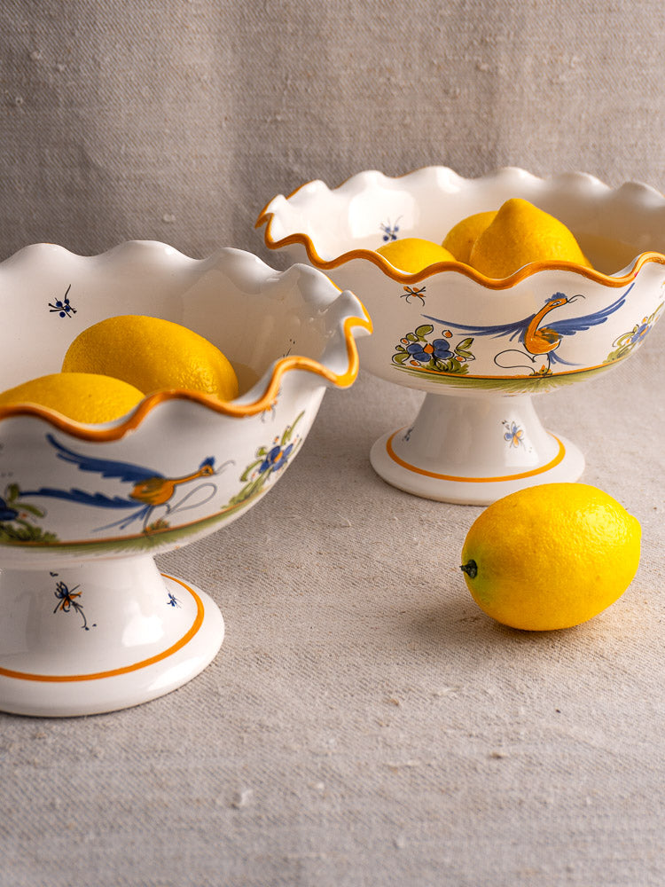 Pair of French Moustiers-style faience compotes with scalloped edges, bird and floral motifs, holding lemons on beige surface.