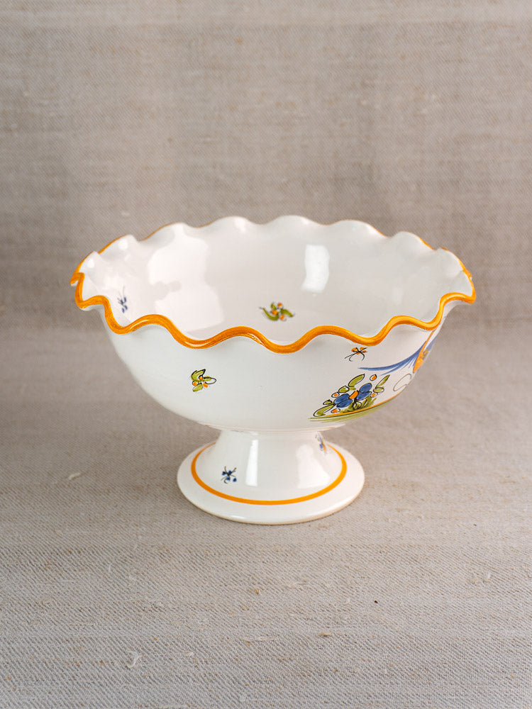Pair of vintage French Moustiers-style faience compotes with scalloped orange-edged rims and floral motifs on fabric background.