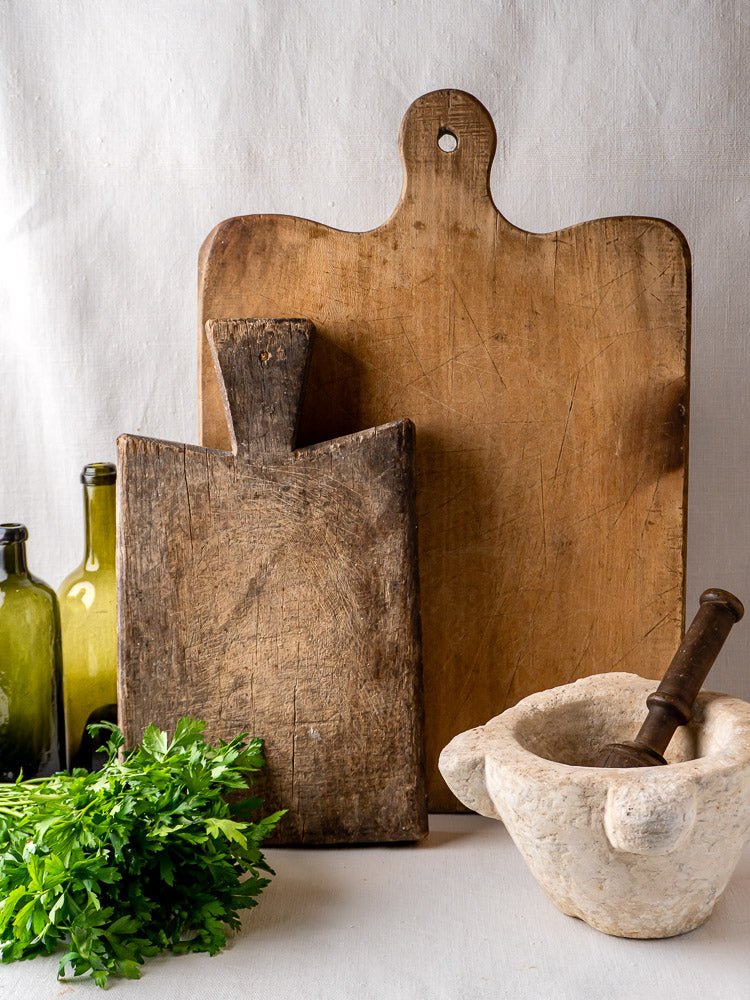 Antique French hardwood chopping board, vintage cutting board, marble mortar with pestle, green glass bottles, and fresh parsley on light cloth.