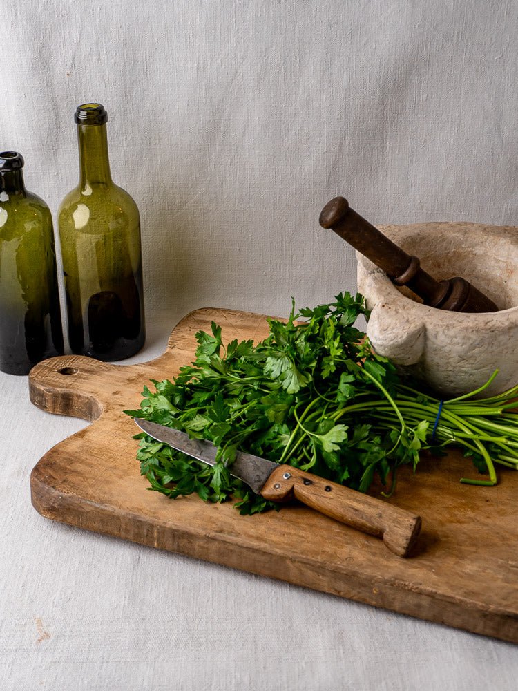 Antique 19th c. French hardwood chopping board with parsley, knife, marble mortar, and green glass bottles on neutral fabric.