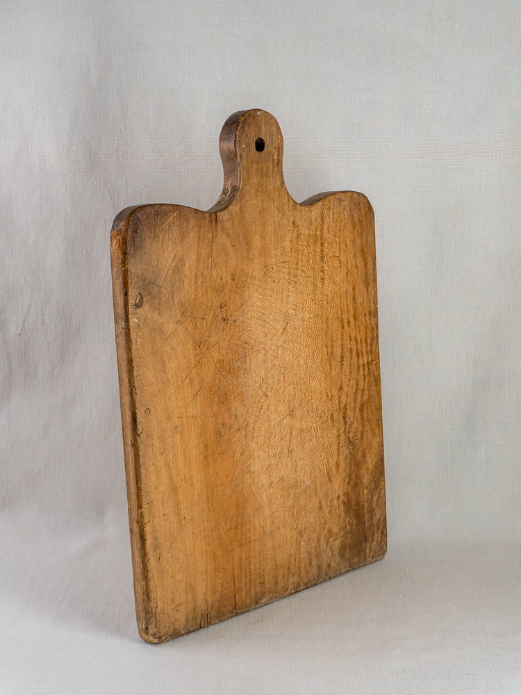 Large 19th-century French hardwood chopping board with rounded handle and hanging hole, against light fabric.