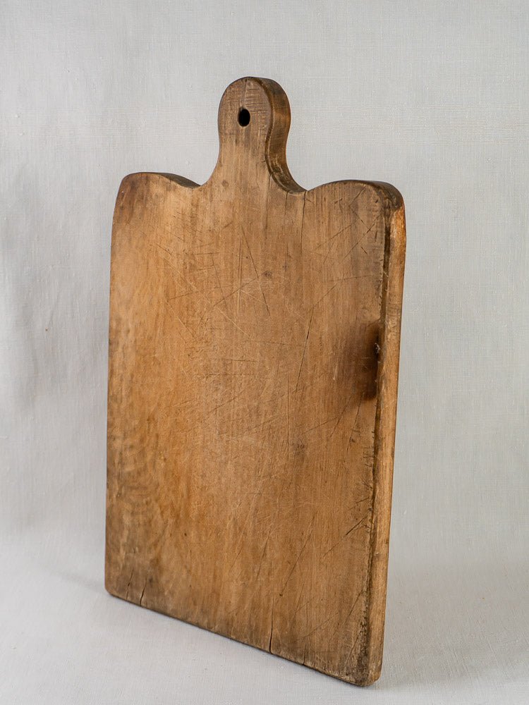 Large 19th-century French hardwood chopping board with rounded top and hanging hole on gray surface.