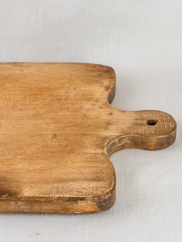 Large antique 19th-century French hardwood chopping board with aged surface and handle, on light gray fabric.