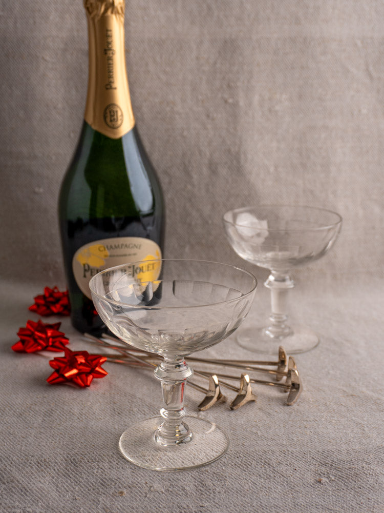 Antique French hand-blown champagne coupes with red gift bows and gold bow-shaped cocktail stirrers on fabric.