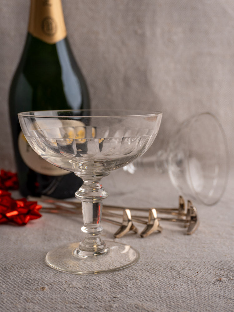 Antique French hand-blown champagne coupes with bottle, metallic stirrers, and a red ribbon on gray textured surface.