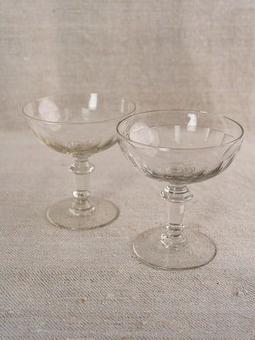 Pair of antique French hand blown champagne coupes with clear glass and short stems on light textured fabric surface.