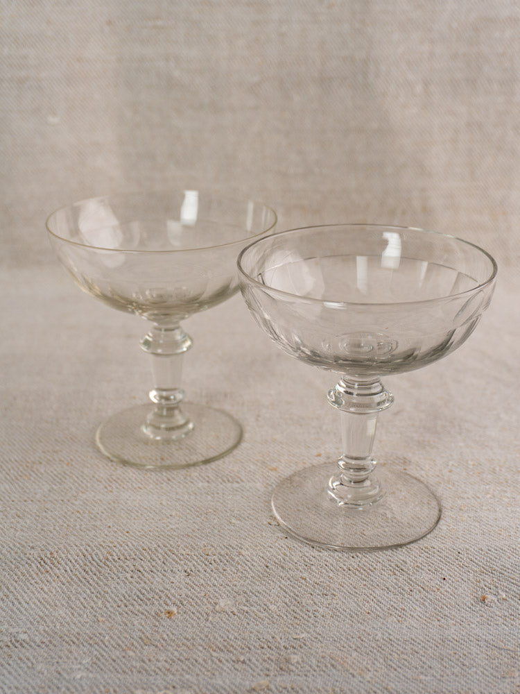 Pair of antique French hand blown champagne coupes with clear glass and short stems on light textured fabric surface.
