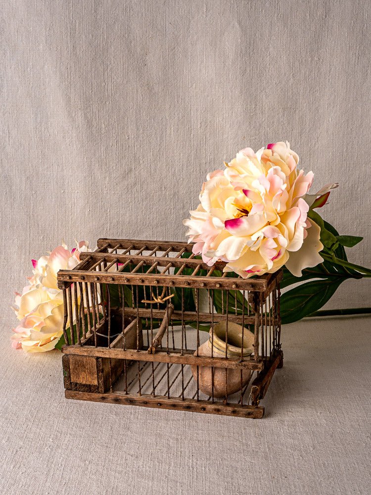 Antique French folk art bird cage in wood and wire with clay pot, perched on neutral fabric amid peonies and green foliage.