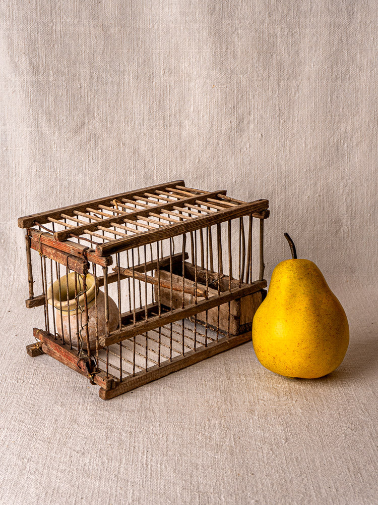 Handmade antique French wood and wire bird cage with vertical bars, perch, and ceramic water container on neutral fabric background.
