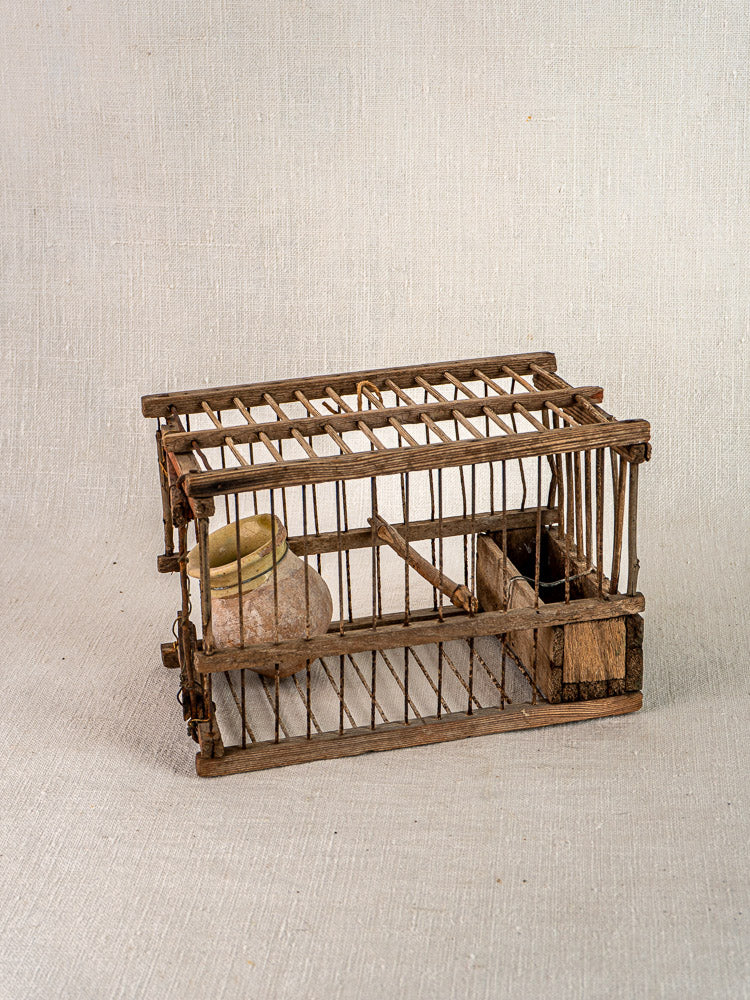 Handmade antique French wood and wire bird cage with vertical bars, perch, and ceramic water container on neutral fabric background.