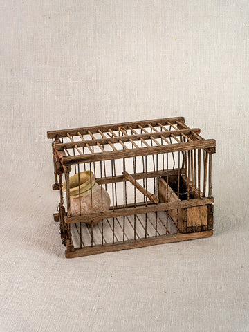 Handmade antique French wood and wire bird cage with vertical bars, perch, and ceramic water container on neutral fabric background.