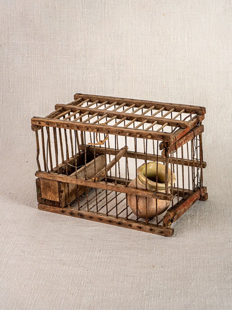 Antique French folk art bird cage in wood and wire with ceramic bowl and wooden feeder on neutral fabric background.