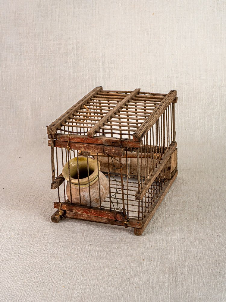 Antique French folk art bird cage in wood and wire with round beige clay pot, displayed on light fabric.