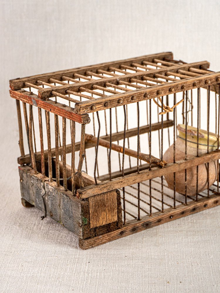 Antique French folk art bird cage in handmade wood and wire, with small clay pot, on light textured surface.