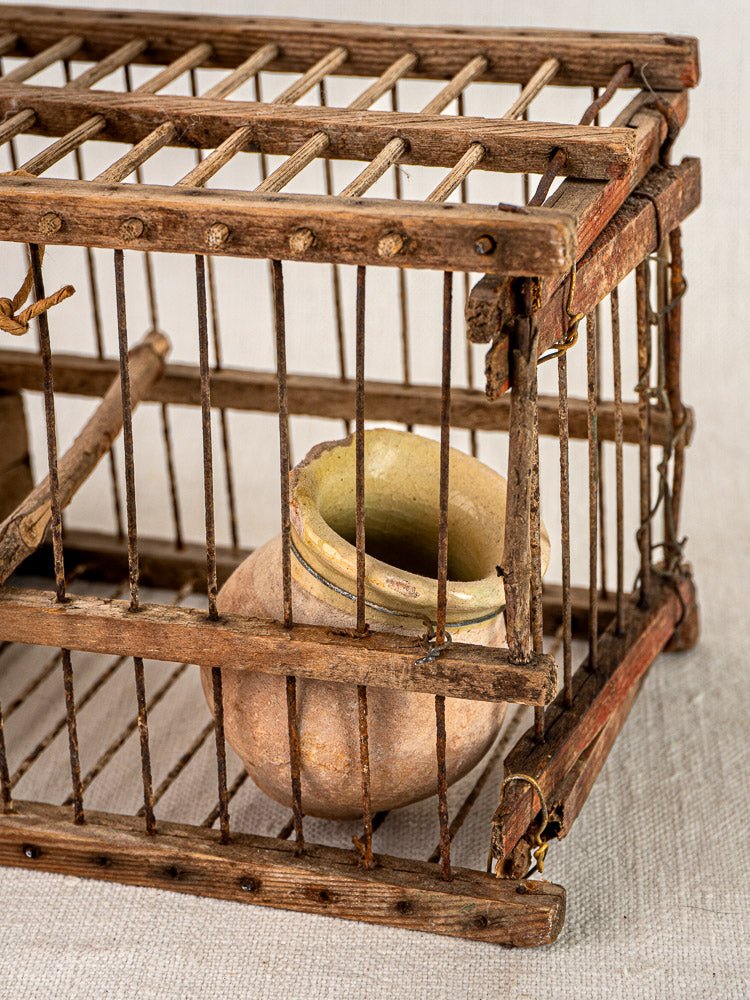 Antique French folk art bird cage, early 20th century wood and wire with ceramic pot, on beige fabric background.