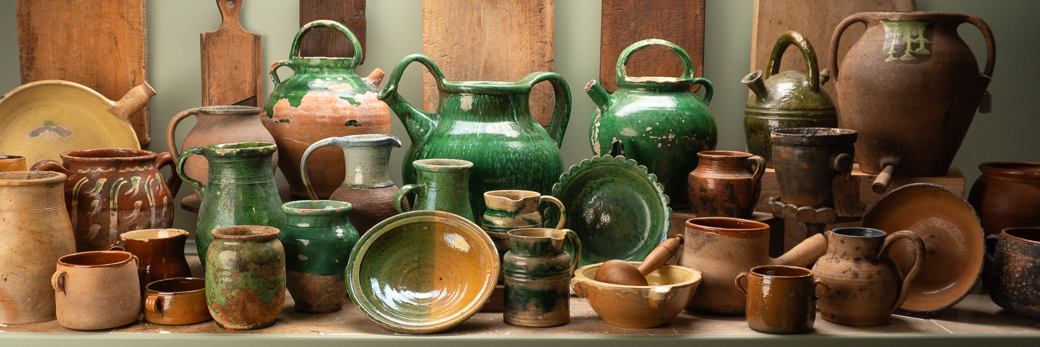 Rustic ceramic jugs, bowls, cups, and pitchers in green and brown on a shelf with wooden backdrop.