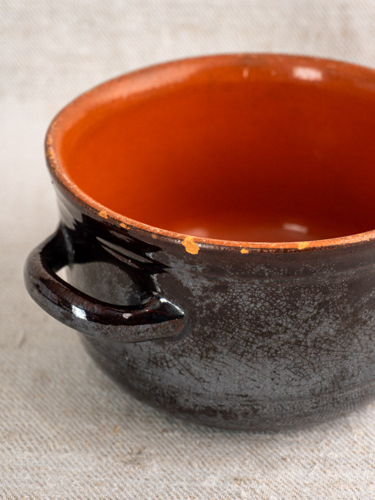 Vintage Italian Albisola earthenware pot, black textured exterior, orange interior, rim chip, side handle, on light fabric.