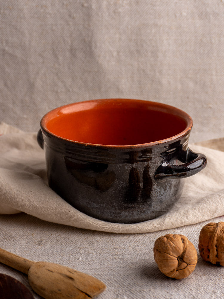 Vintage Albisola glazed earthenware cooking pot with black exterior, orange interior, displayed with walnuts and wooden spoon.