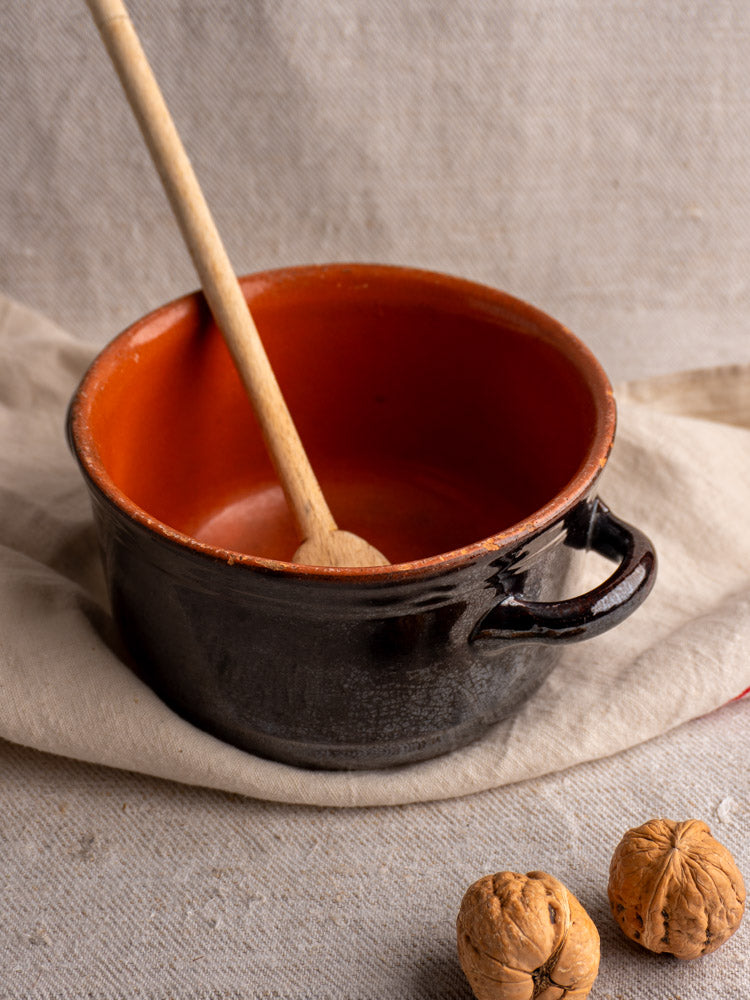 Albisola glazed earthenware cooking pot with black exterior, reddish-brown interior, and wooden spoon on beige cloth.