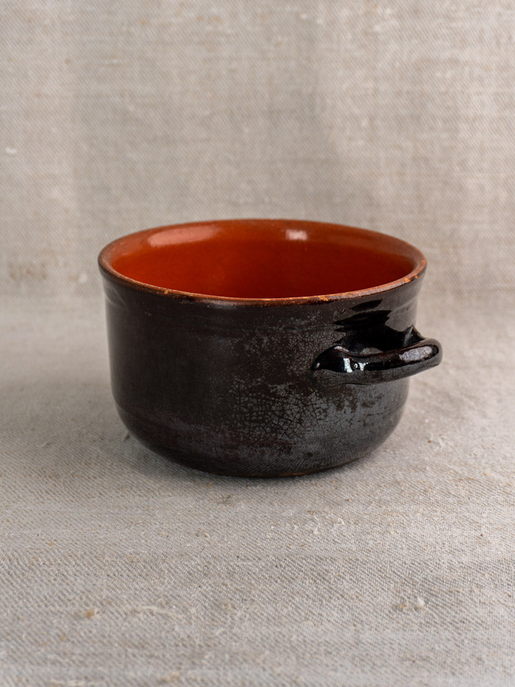 Vintage Albisola glazed earthenware cooking pot, dark brown with orange interior, single handle on fabric background.