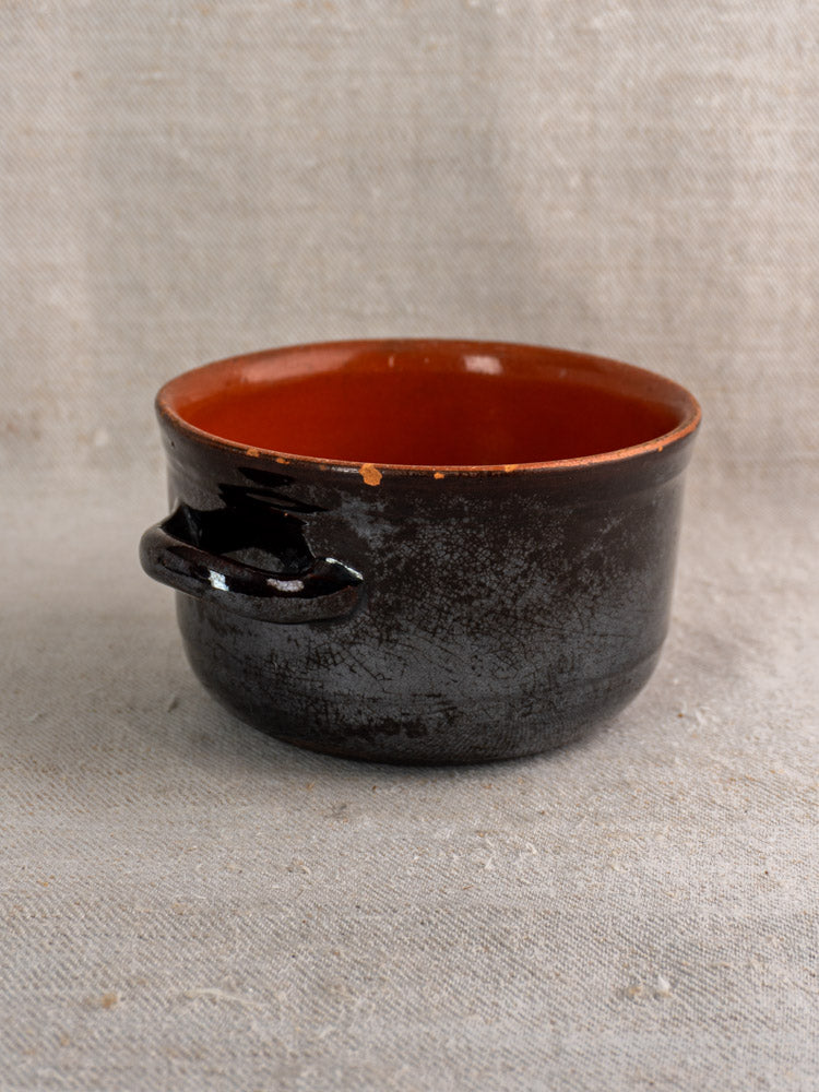 Vintage Albisola black glazed earthenware cooking pot with small handle and reddish-brown interior on light fabric.