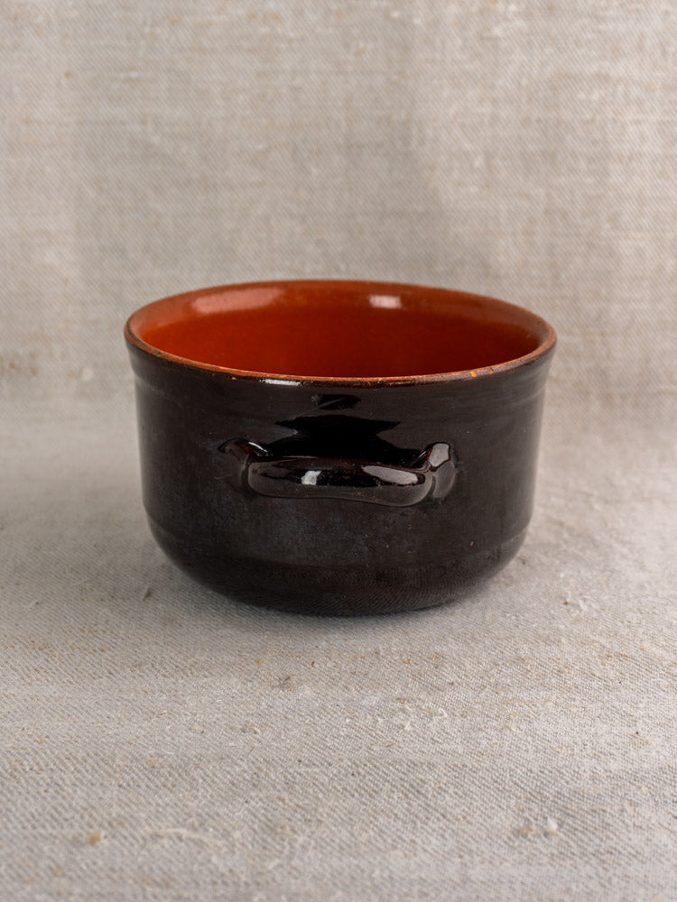 Small Albisola glazed earthenware Italian cooking pot, glossy dark brown with side handle, on textured beige fabric.