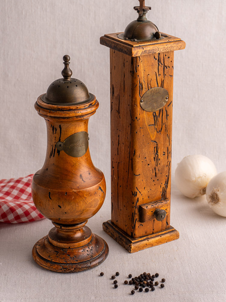 Antique Italian wormwood pepper mill and salt set with brass accents on linen, surrounded by whole peppercorns and onions.