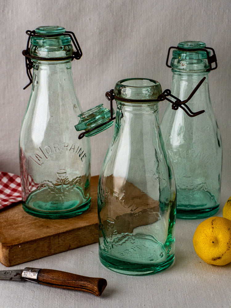 Set of three antique French La Lorraine aqua glass preserving bottles with lemons and red-checked cloth.